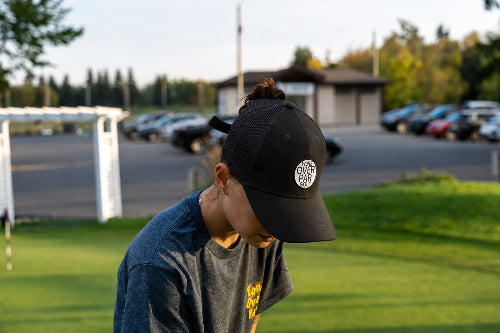 A person outdoors on a grassy field wearing a black pony tail cap with a circular white logo and a dark gray t-shirt, with a blurred background of parking lot, trees, and a building.