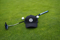 A black Pony Tail-style cap with a circular 'Fore Over Par' logo lies on a green grass surface, crossed by a  golf putter and flanked by two white golf balls.