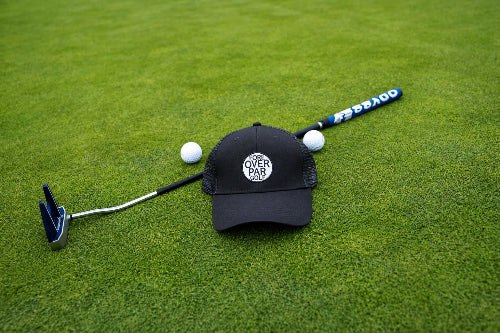 A black Pony Tail-style cap with a circular 'Fore Over Par' logo lies on a green grass surface, crossed by a  golf putter and flanked by two white golf balls.