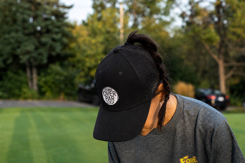 A 5'1" Woman outdoors on a grassy field wearing a black pony tail-style  baseball cap with a circular white and black 'Fore Over Par' patch on the front, along with a gray t-shirt.