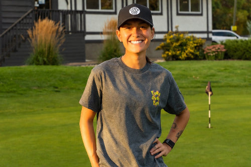 A 5'1" woman stands on a golf course wearing a gray t-shirt with a yellow logo,  and a black pony tail cap. She is holding a golf club, with a modern house and green grass in the background.