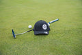 A black baseball cap with a circular white and black logo on the front, placed on a green grass surface next to a blue and white golf putter, a white golf ball, and another golf ball.