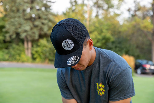 A man outdoors on a grassy field wearing a black trucker-style baseball cap with a circular sticker on the brim and a dark gray t-shirt with a yellow graphic.