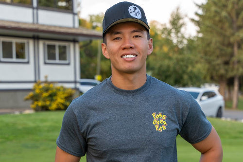 A 5'9" smiling man standing on a golf course wearing a dark gray heathered t-shirt with a yellow embroidered logo on the left chest, a dark baseball cap, and dark pants.