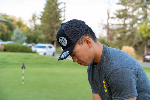 A man wearing a black trucker-style baseball cap with a circular sticker and a gray t-shirt, looking down on a golf course green.
