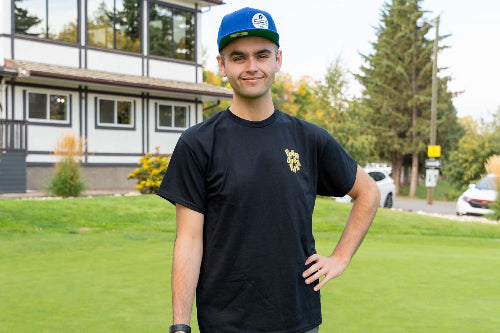 A 6'1"man standing on a golf course wearing a large black t-shirt with a small gold crest on the left chest, gray shorts, a blue cap, and holding a golf club.