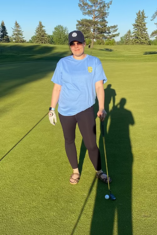 5'5" Female walking on a golf course holding a golf club  wearing a blue Fringe Society Tee size large and Dark Blue Pony Tail Club Hat by Fore Over Par with a clear blue sky and trees in the background.