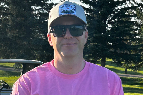 A man wearing a pink t-shirt, black sunglasses, and a sand baseball cap with a 'Fore Over Par' patch, standing outdoors on a golf course.