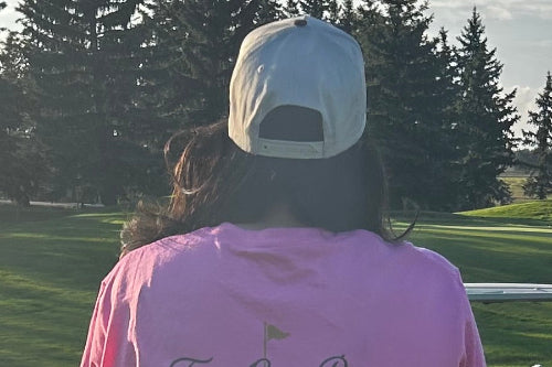 A person viewed from behind wearing a sand baseball cap and a pink t-shirt, standing on a golf course with trees and grass in the background.