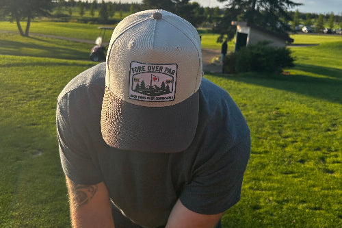 A man on a golf course wearing a sand and brown baseball cap with a 'Fore Over Par' patch on the front, looking down while wearing a dark gray t-shirt.