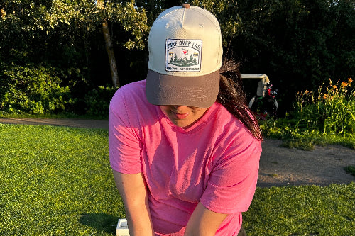 A person on a golf course wearing a pink t-shirt, and a beige and brown trucker hat with a 'Fore Over Par' logo, holding a golf club.