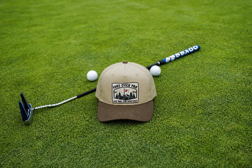 A beige and brown baseball cap with a 'Fore Over Par' graphic patch, styled on a green grass field with a golf putter, golf balls, and a golf club.