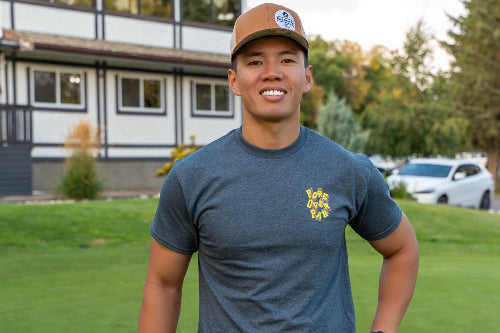 A 5'10" man standing on a golf course wearing a dark gray t-shirt with a yellow logo on the left chest, dark pants, and a brown baseball cap, holding a golf club.