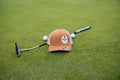 A caramel & khaki baseball cap with a circular 'The Game' logo on the front, placed on a green golf course with a putter, golf balls, and a golf club.