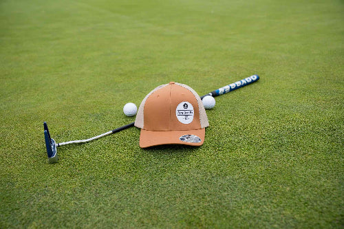 A caramel & khaki baseball cap with a circular 'The Game' logo on the front, placed on a green golf course with a putter, golf balls, and a golf club.