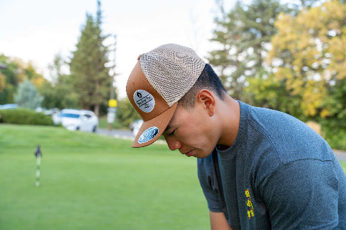 A man on a golf course wearing a tan and beige trucker hat with a circular logo patch on the front, paired with a dark gray t-shirt.