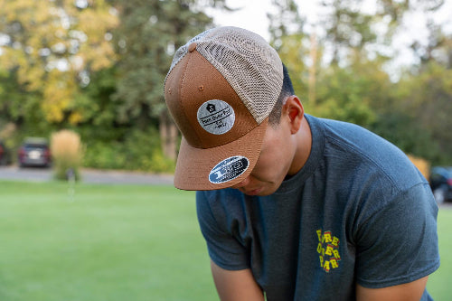 A man wearing a brown and beige trucker hat with a circular 'Brewery Select' logo on the front, paired with a dark blue t-shirt, looking down in an outdoor setting with a blurred green lawn and trees in the background.