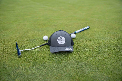 A Charcoal and Black OG hat by Fore Over Par shown on a golf green with a putter and two golf balls behind it.