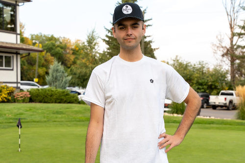 A 6'1" man standing on a golf course wearing a white crewneck t-shirt with a small 'Fore Over Par' Logo on the left chest, gray shorts, and a black baseball cap, holding a golf club.
