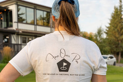 A 5'10" woman on a golf course seen from behind, wearing a white graphic t-shirt with the text 'Fore Over Par, for the love of the game, not the score' and a navy blue skirt.