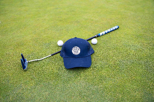Dark Blue Pony Tail Club Hat by Fore Over Par, shown on golf green with putter and golf balls behind it. 