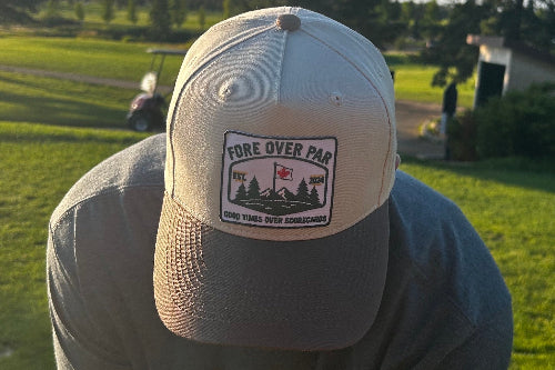 A man on a golf course wearing a sand coloured, green brimmed baseball cap with a front patch that reads 'FORE OVER PAR' and 'GOOD TIMES OVER SCORECARDS'