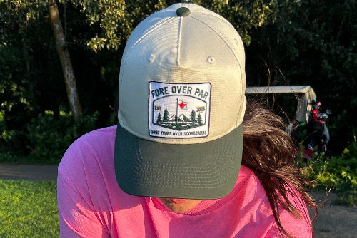 A woman on a golf course wearing a sand and green snapback cap with a 'FORE OVER PAR' graphic patch on the front, paired with a bright pink t-shirt.