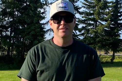 A man wearing a sand trucker hat with  a green brim, and a 'Fore Over Par' logo patch, black sunglasses, and a dark green t-shirt, standing on a golf course in front of trees and grass.