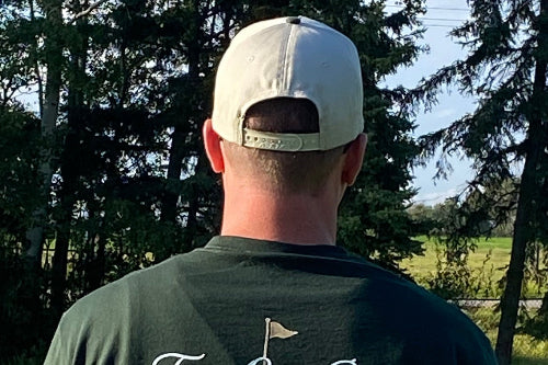 A man viewed from behind wearing a sand baseball cap and a dark green t-shirt with a golf flag graphic on the back, standing on a golf course in front of trees.