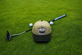 A sand and green baseball cap with a 'Fore Over Par' graphic patch lies on a green grass surface, styled with a golf putter, and two white golf balls.