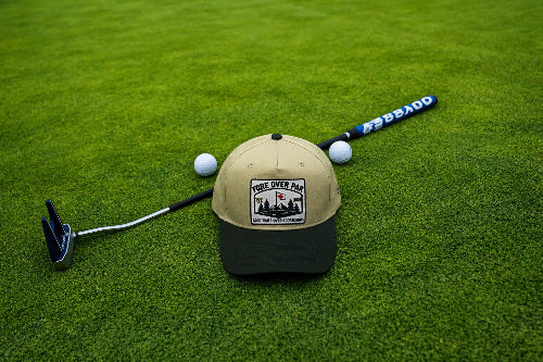A sand and green baseball cap with a 'Fore Over Par' graphic patch lies on a green grass surface, styled with a golf putter, and two white golf balls.