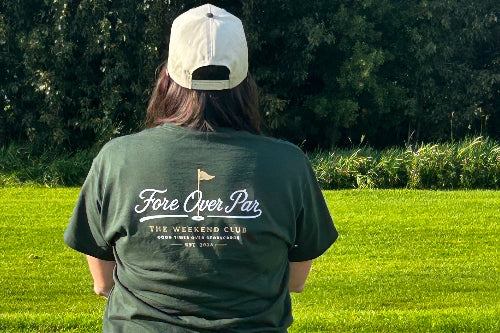 A person standing on a grassy golf course, viewed from behind, wearing a dark green t-shirt with 'Fore Over Par' printed on the back, a black pleated skirt, and a sand cap.