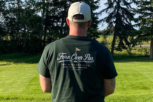 A man standing outdoors on a golf course with trees in the background, viewed from behind. He is wearing a dark green t-shirt with 'Fore Over Par' and 'The Weekend Club' printed on the back, along with a light-colored baseball cap.