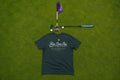 A green t-shirt laid flat on a green grass surface, with a golf putter and flagstick placed above it. The shirt has a white script logo reading 'Fore Over Par' and smaller text below.
