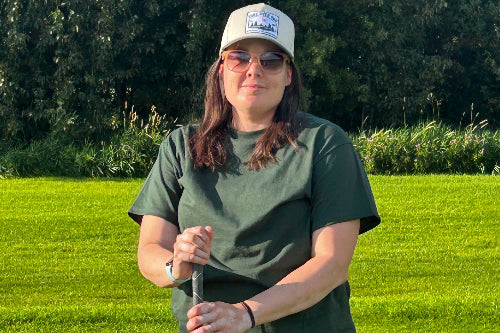 A 5'5" woman standing on a golf course while wearing a green t shirt, holding a golf club, and wearing sand coloured hat. 