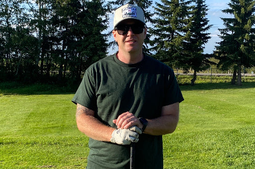 A 5'9" man on a golf green wearing a green t-shirt while holding a golf club and wearing a sand coloured hat. 