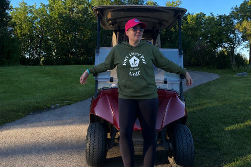A 5'5" woman stands on a paved path in front of a red and white golf cart, wearing a green hoodie with white 'Fore Over Par Golf' Logo, black leggings, and a pink cap. Trees and grass are visible in the background under a clear blue sky.