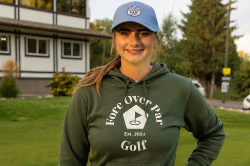 A 5'10" woman stands on a golf course wearing an green hoodie with 'Fore  Over Par Golf' printed on the front, a blue cap, and a navy blue skirt, holding a golf club.