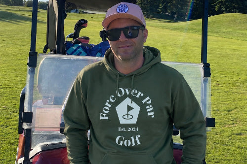 A 5'9" man wearing a green 'Fore Over Par Golf' hoodie,  and a pink cap stands in front of a red golf cart on a sunny golf course.