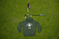 A green pullover hoodie with a white 'Fore Over Par Golf' graphic is laid flat on a green grass lawn, styled with a miniature golf hole setup (flag, tee, club, and golf balls) above it.
