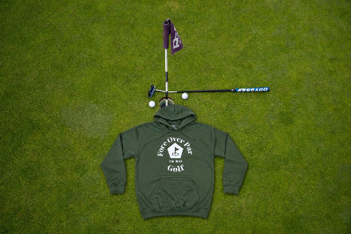 A green pullover hoodie with a white 'Fore Over Par Golf' graphic is laid flat on a green grass lawn, styled with a miniature golf hole setup (flag, tee, club, and golf balls) above it.