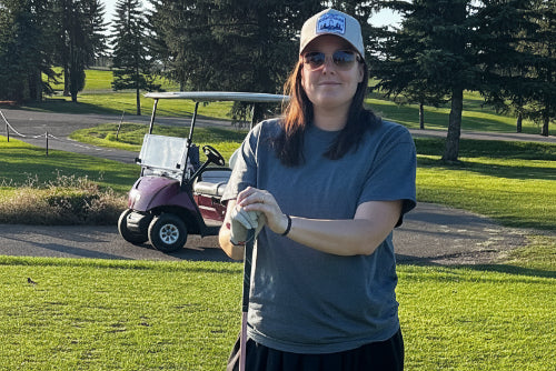 A 5'5" woman on a golf course wearing a grey t-shirt, black skirt, and a baseball cap, holding a golf club with a golf ball in front of her. A golf cart is visible in the background.