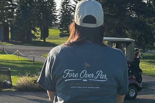 A 5'5"woman standing on a golf course, viewed from behind, wearing a grey t-shirt with 'Fore Over Par' printed on the back, a white cap, and a black skirt. A golf cart is visible in the background on the grass.