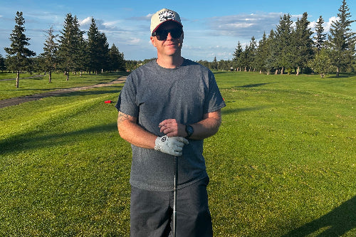 A 5'10" man wearing a grey t-shirt, dark grey shorts, a white cap, and sunglasses stands on a golf course holding a golf club under a clear blue sky.