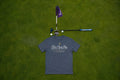A grey  t-shirt with a 'Fore Over Par' graphic on the back , laid flat on a green grass background next to a miniature golf hole setup with a purple flag, club, and golf balls.