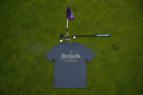 A grey  t-shirt with a 'Fore Over Par' graphic on the back , laid flat on a green grass background next to a miniature golf hole setup with a purple flag, club, and golf balls.