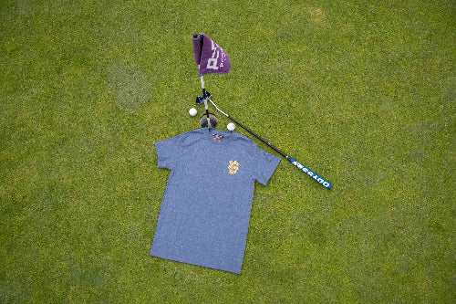 A grey short-sleeve t-shirt with a small colourful crest 'Fore Over Par' logo on the left chest, laid flat on a green grass surface next to a purple flag, golf balls, and a golf club.