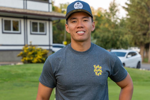 A 5'10" man wearing a grey t-shirt with a colourful 'Fore Over Par logo on the left chest, a navy blue cap, and dark pants, standing on a golf course holding a club.