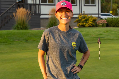 A 5'1" woman on a golf course wearing a grey t-shirt with a colourful 'Fore Over Par' Logo, with a house in the background.