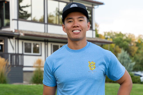 A 5'10" Man in a large light blue shirt and black pants standing on a golf course with a building in the background